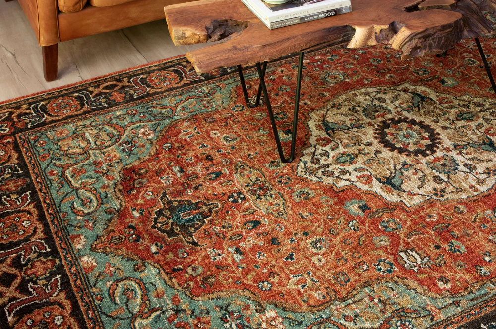 Oriental-style rug with a central medallion and intricate floral motifs in red, teal, cream, and black, paired with a live-edge wooden coffee table holding books, and a tan leather armchair partially visible in the corner.