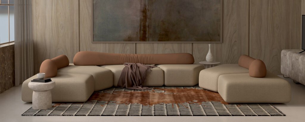 Modern minimalist living room with modular beige sectional sofa featuring rounded edges and brown cylindrical backrests, brown throw blanket, patterned area rug in brown and black tones, stone side table with books and cup, abstract earthy-toned painting on wood-paneled wall, and natural light through sheer curtains