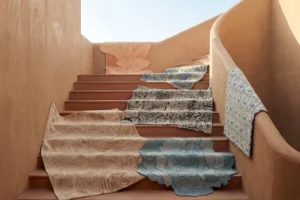 Indian carpets, intricately designed rugs with unique patterns and textures including peach leaf motifs, blue floral designs, black and white abstracts, and embossed styles, draped over a staircase to showcase traditional craftsmanship and modern artistry
